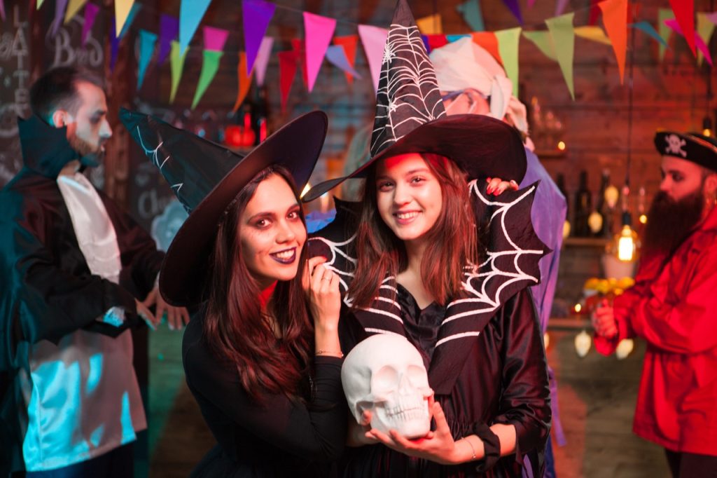 Beautiful young witch holding a human skull and smiling to the camera at halloween party. Witches looking into the camera.