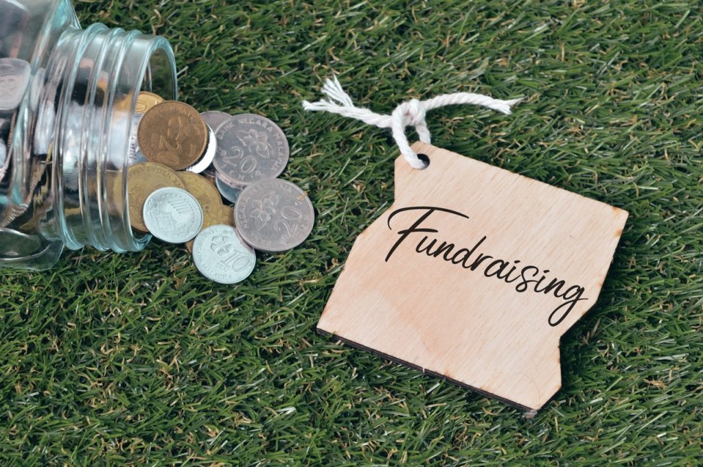 A glass jar tipped over on a green grass lawn, spilling out various coins next to a small, rustic wooden tag with the word "Fundraising" written on it in black cursive script.